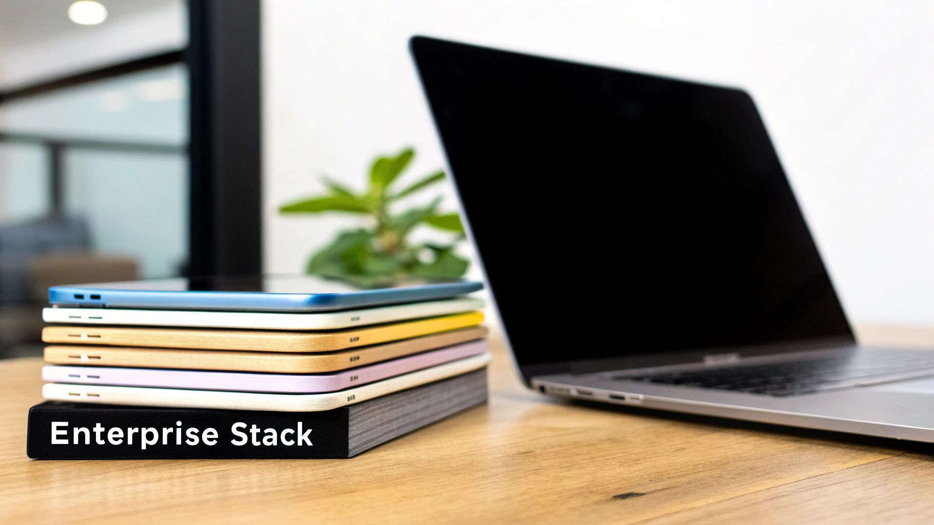 A stack of multi-colored tablets on an 'Enterprise Stack' book next to a laptop on a wooden desk.