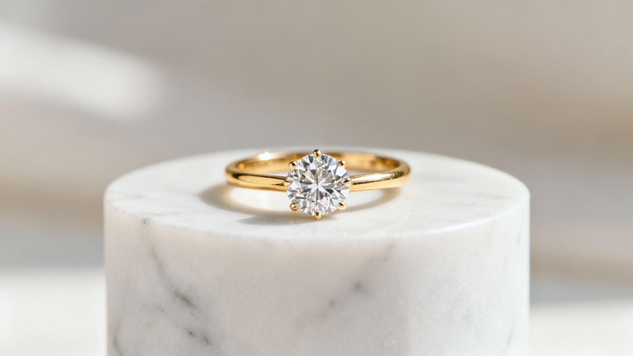 טבעת אירוסין מזהב יוקרתית עם יהלום עגול מנצנץ, מונחת על הדום שיש לבן, בתאורה רכה.