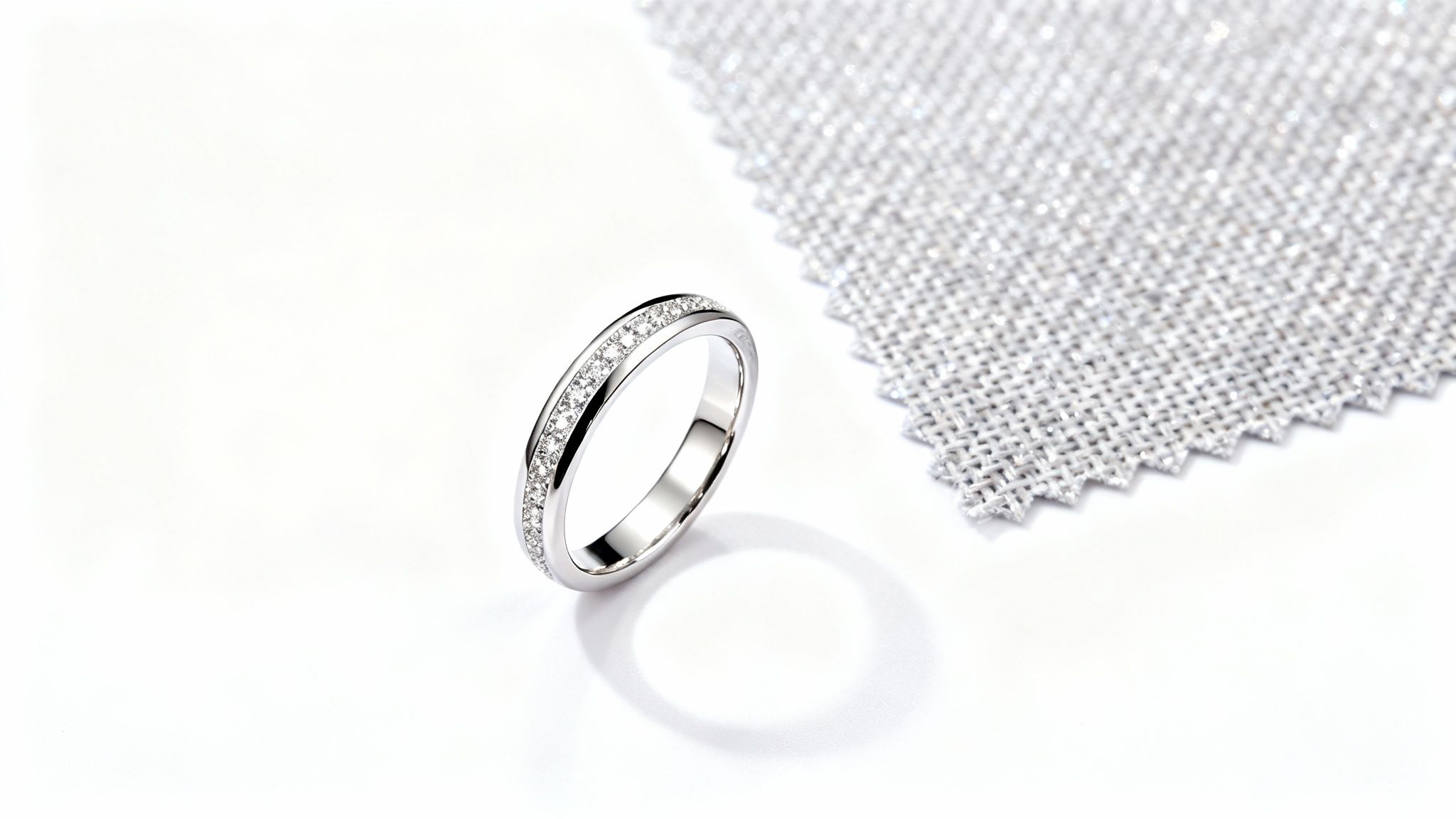 טבעת כסף נוצצת משובצת יהלומים מבריקים על רקע לבן, לצד פיסת בד כסופה ונוצצת.