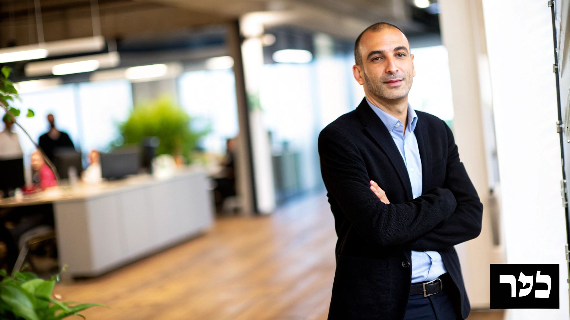 גבר מחייך בחולצה כחולה וז'קט כהה עומד עם ידיים שלובות במשרד מודרני מואר.