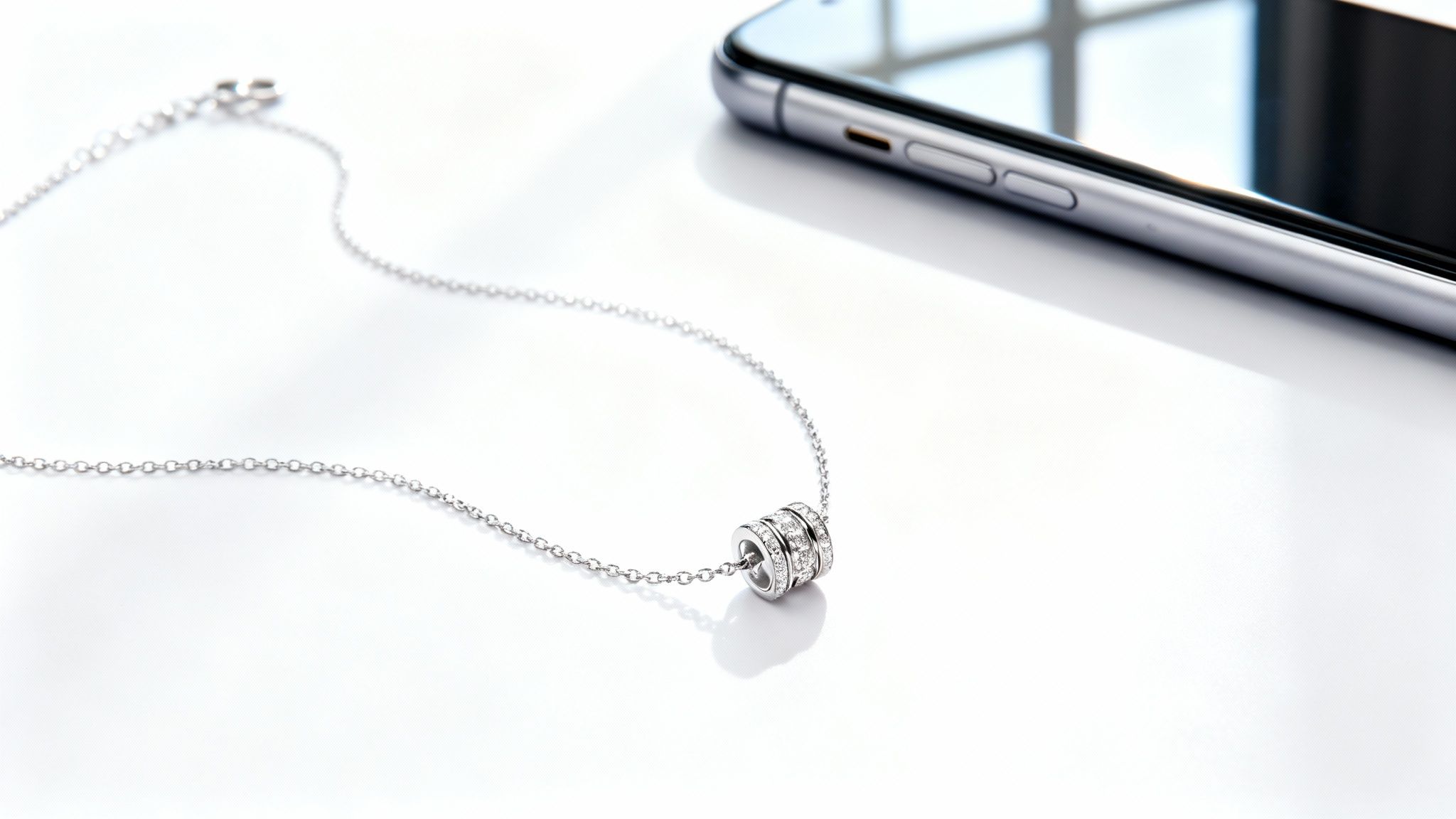 שרשרת כסף עדינה עם תליון גלילי משובץ יהלומים, מונחת על רקע לבן ליד טלפון סלולרי.