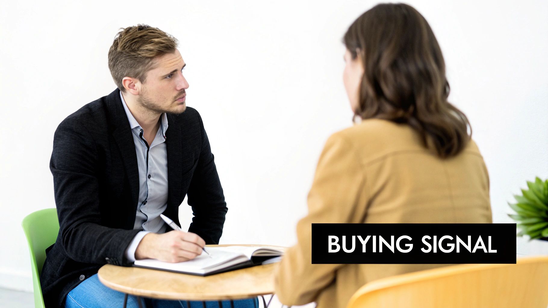 A man in a black blazer writes in a notebook, looking at a woman during a sales meeting, with 'BUYING SIGNAL' text overlay.