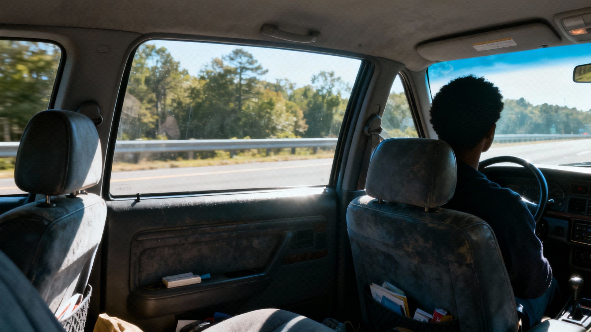 Rear view of a person with afro hair driving a car on a sunny highway, seen from inside the back seat.