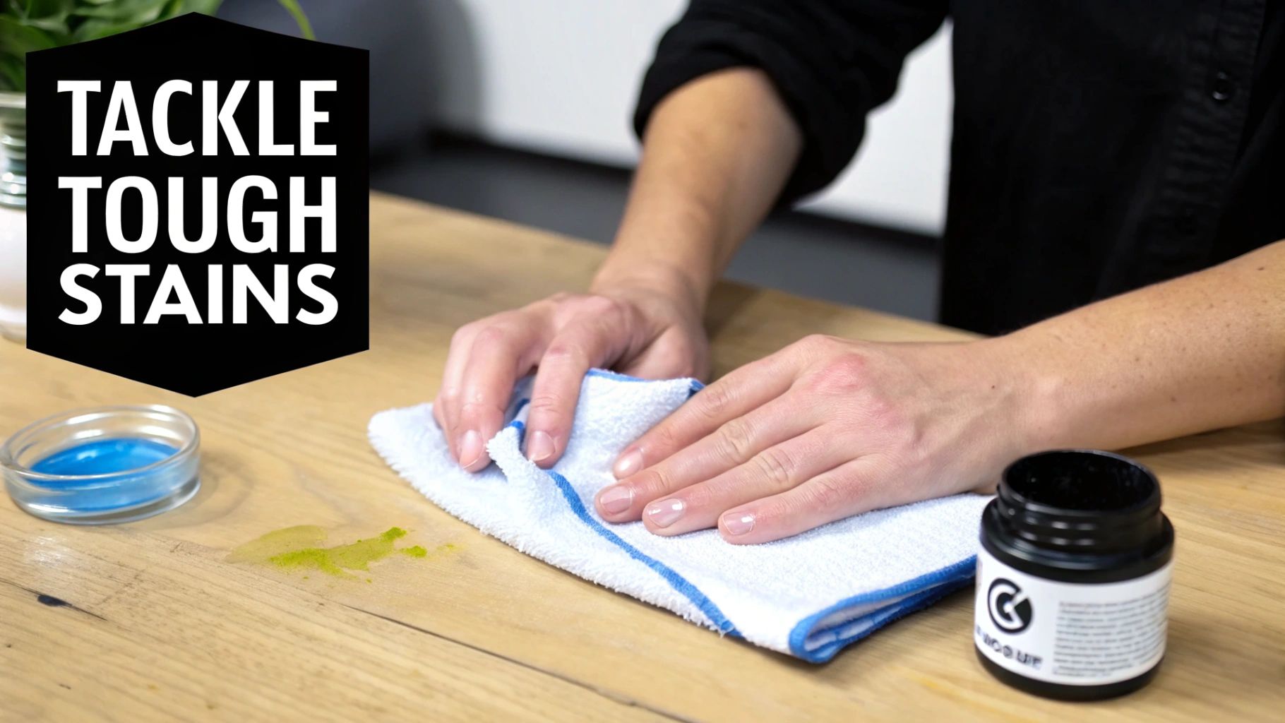 Hands using a white cloth to clean a green stain from a wooden table, next to cleaning products.
