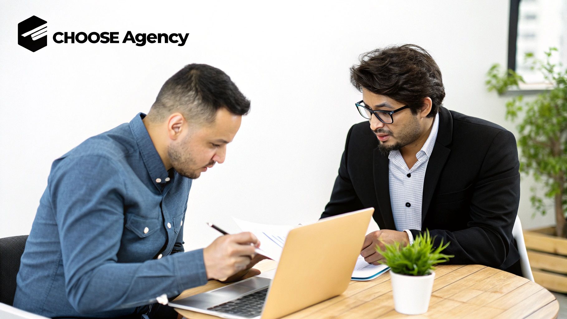 Two business professionals discussing documents and working on a laptop at a wooden desk.