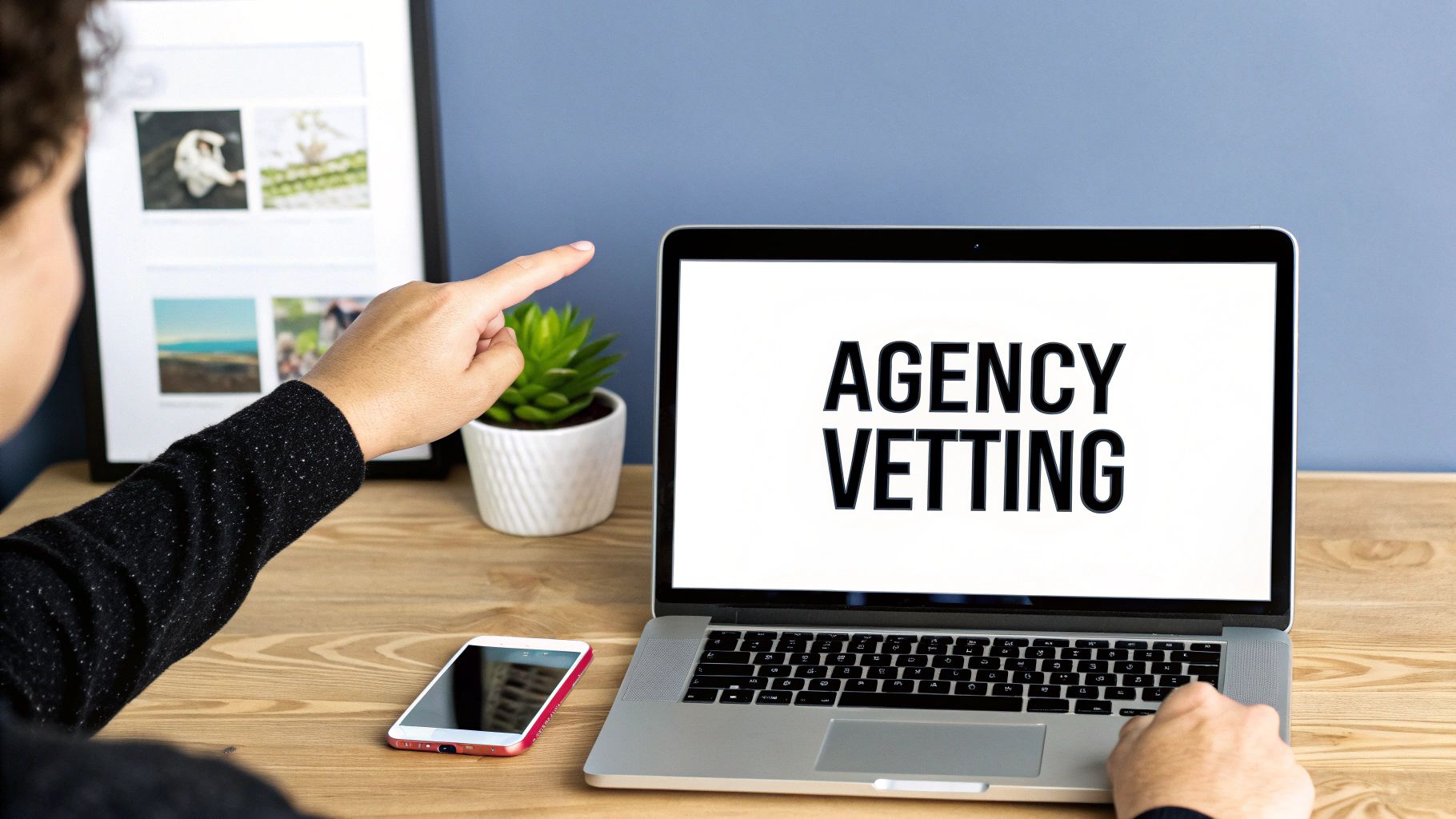 A person's hand points at a laptop displaying 'AGENCY VETTING' on a wooden desk, with a phone nearby.