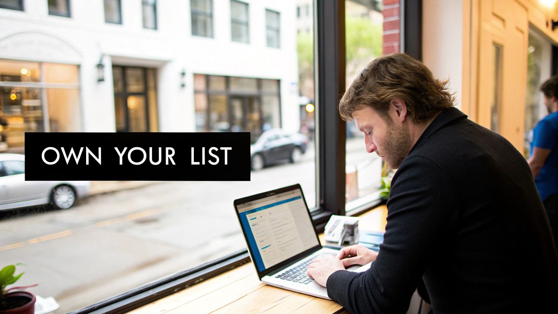 A man with a beard works on a laptop by a window, with a street view and 'OWN YOUR LIST' text.