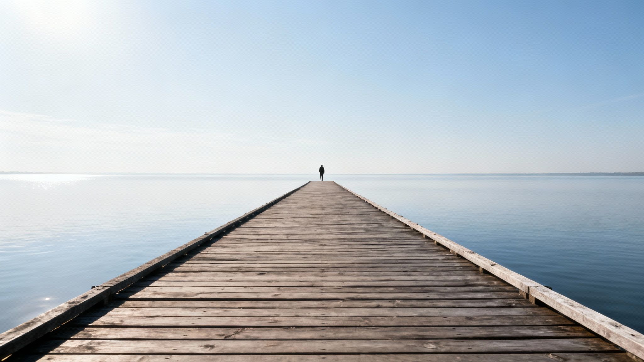 A lone person stands at the end of a long wooden pier stretching into calm, reflective water under a clear sky.