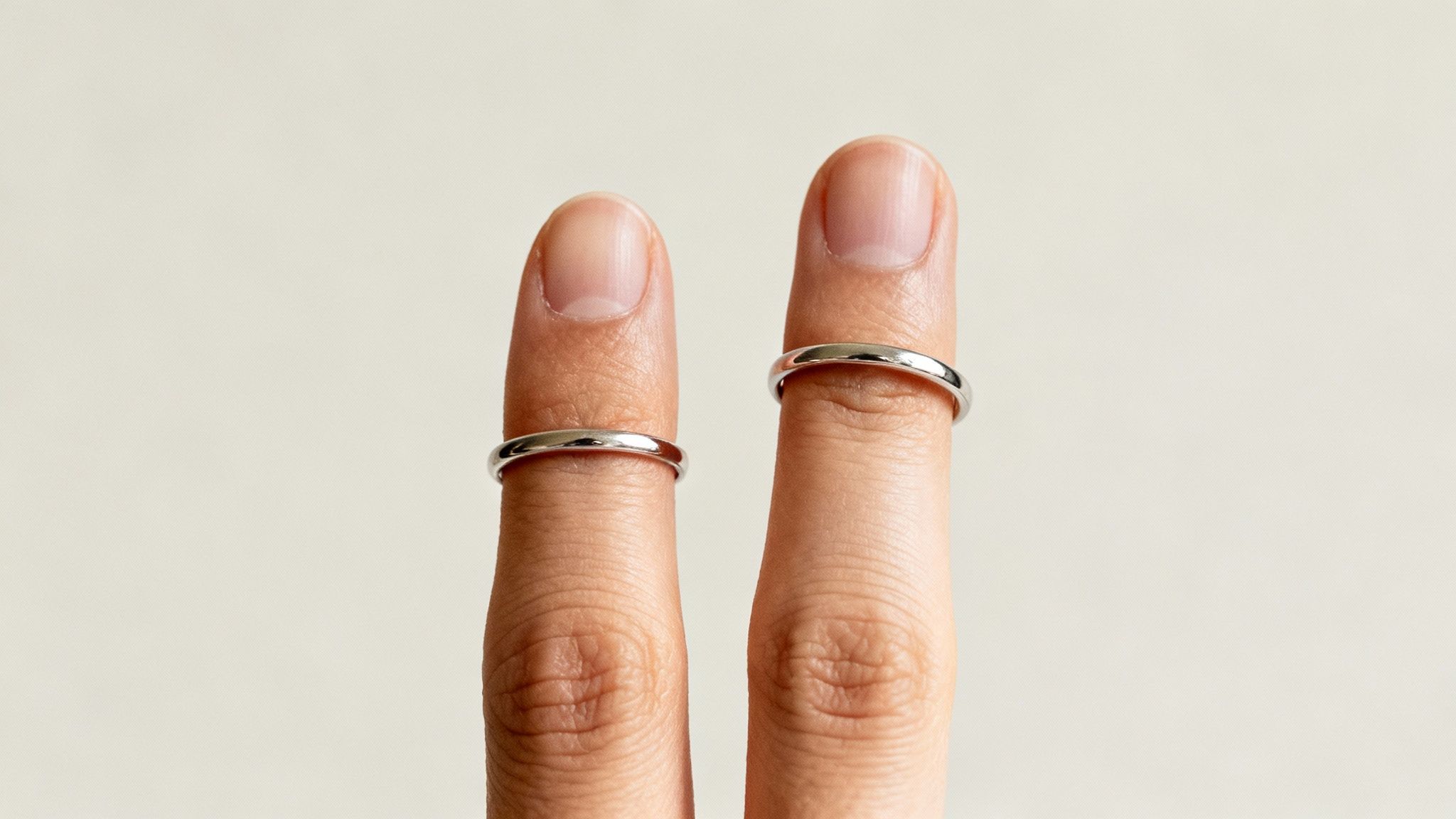 Close-up of two fingers, each wearing a plain silver band ring, against a light background.