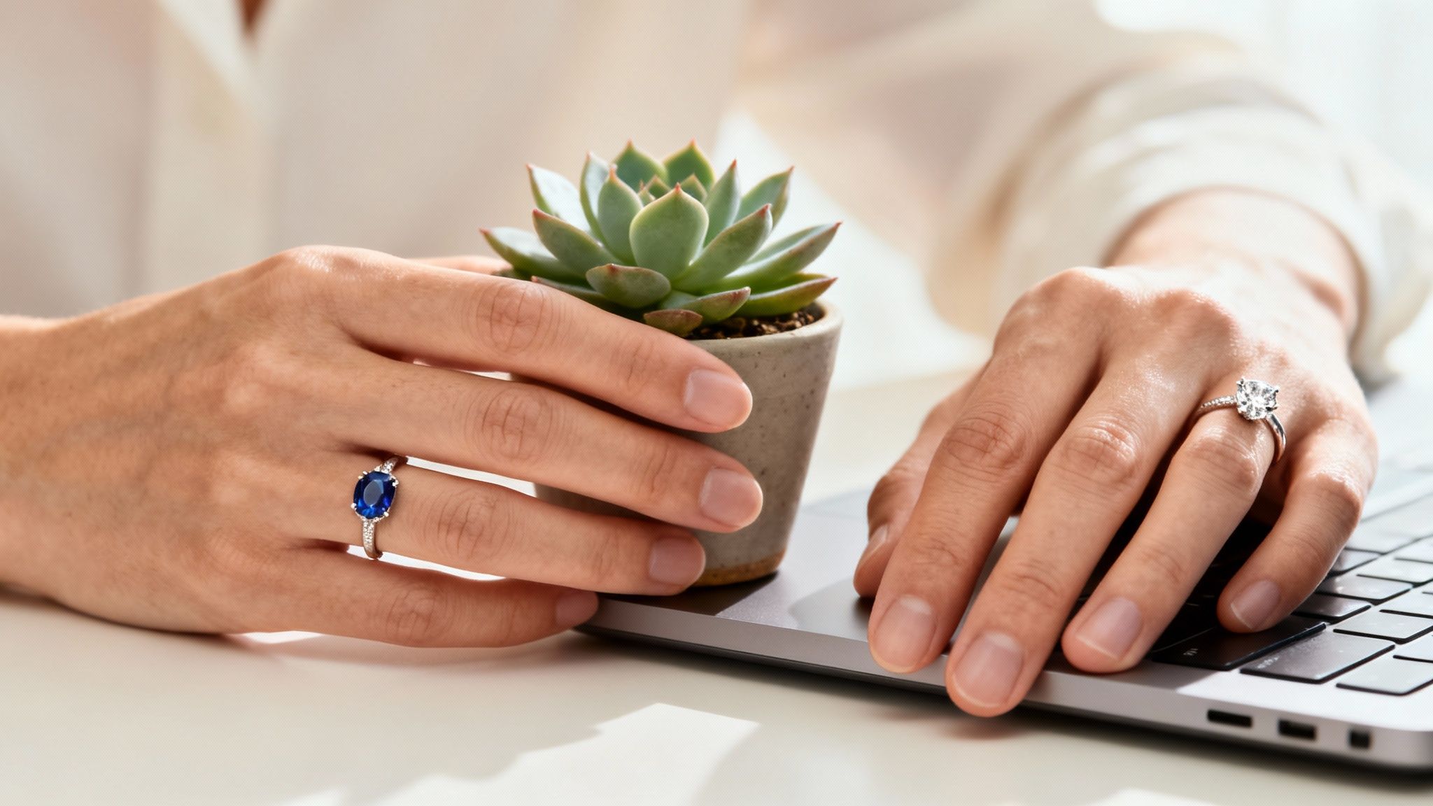 Hands wearing sapphire and diamond engagement rings, holding a succulent plant at a laptop.