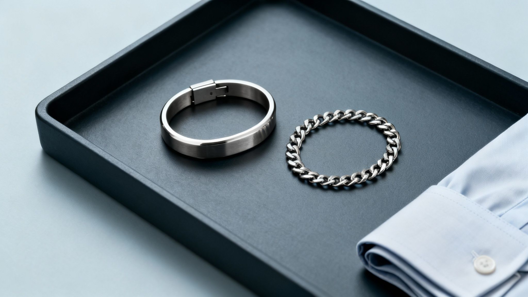 Two stylish silver bracelets, a bangle and a chain, on a dark tray next to a blue shirt cuff.
