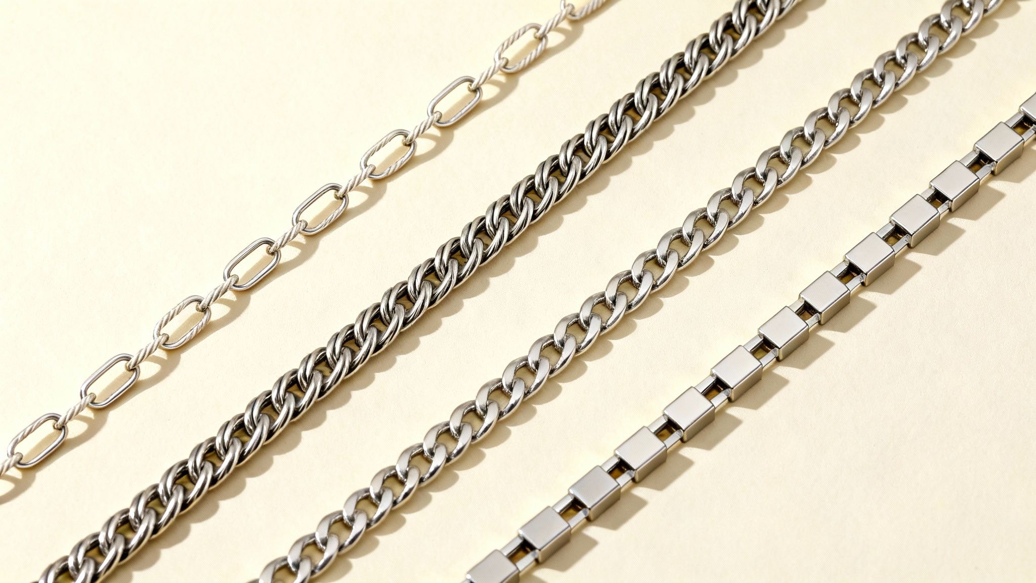 Four silver-toned metal chains of various designs, including twisted oval, Cuban, and rectangular links, on a beige surface.