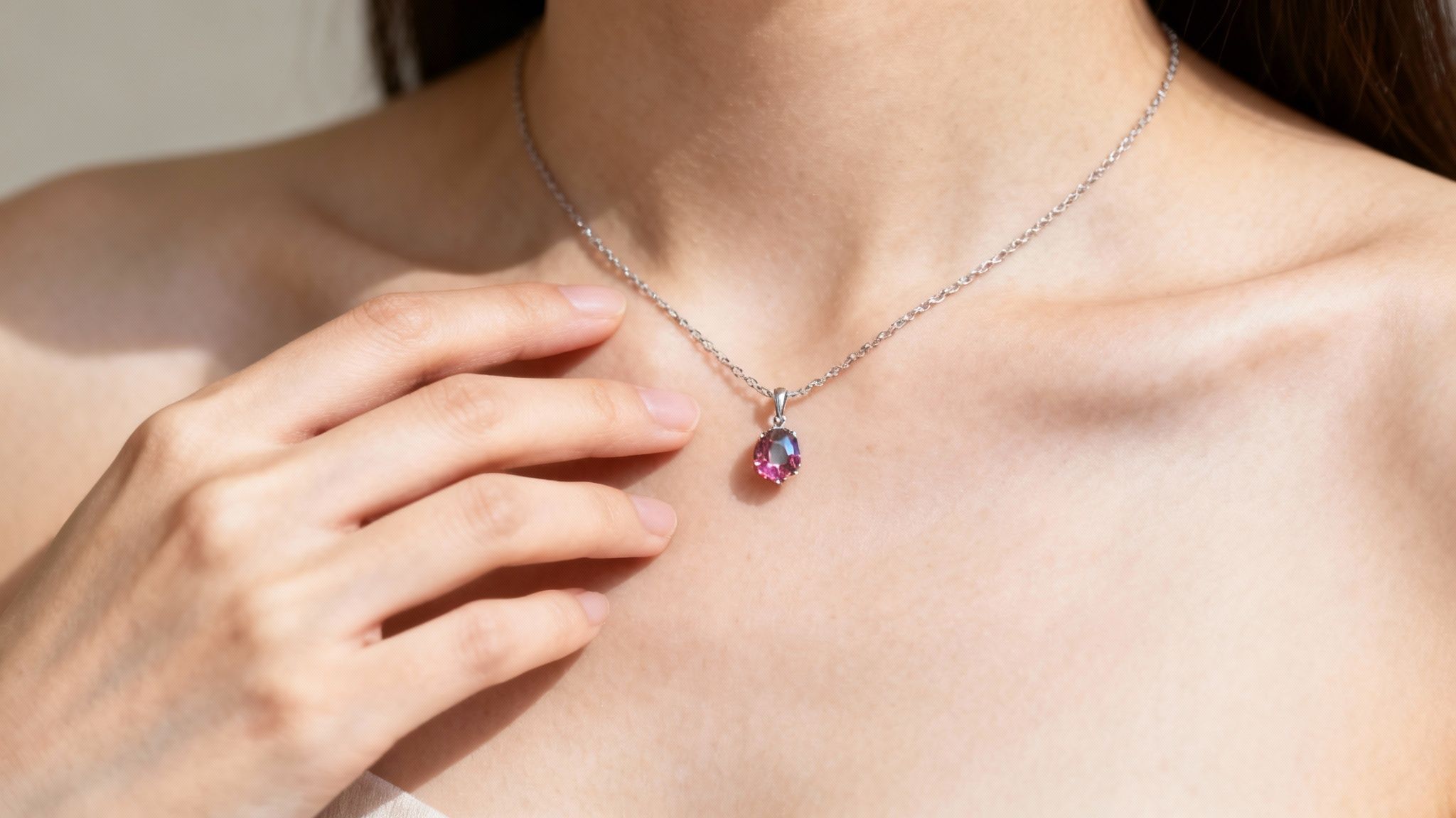 Close-up of a person's chest with a silver necklace featuring a sparkling purple-pink gemstone pendant, touched by a hand.