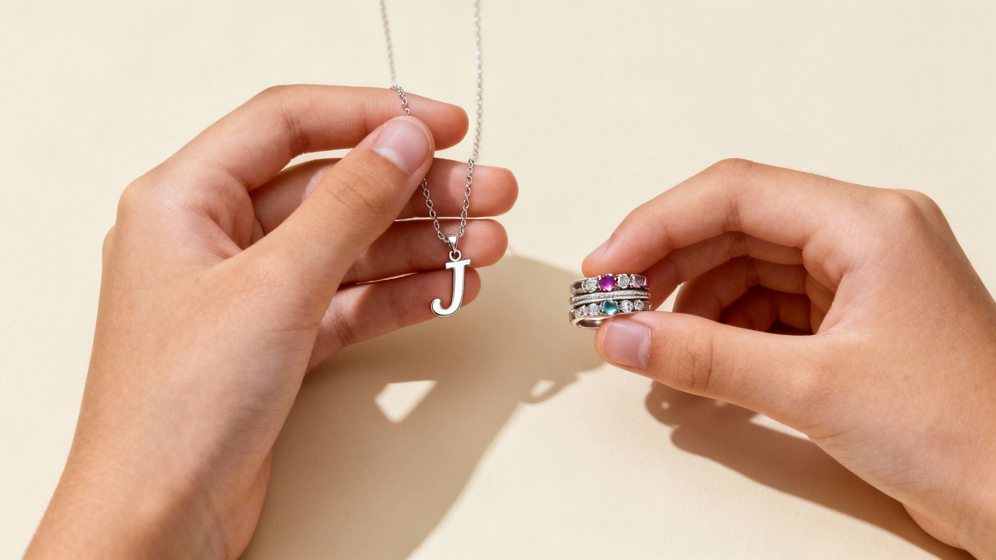 Close-up of hands holding a silver 'J' initial necklace and stacked rings with colorful gemstones.