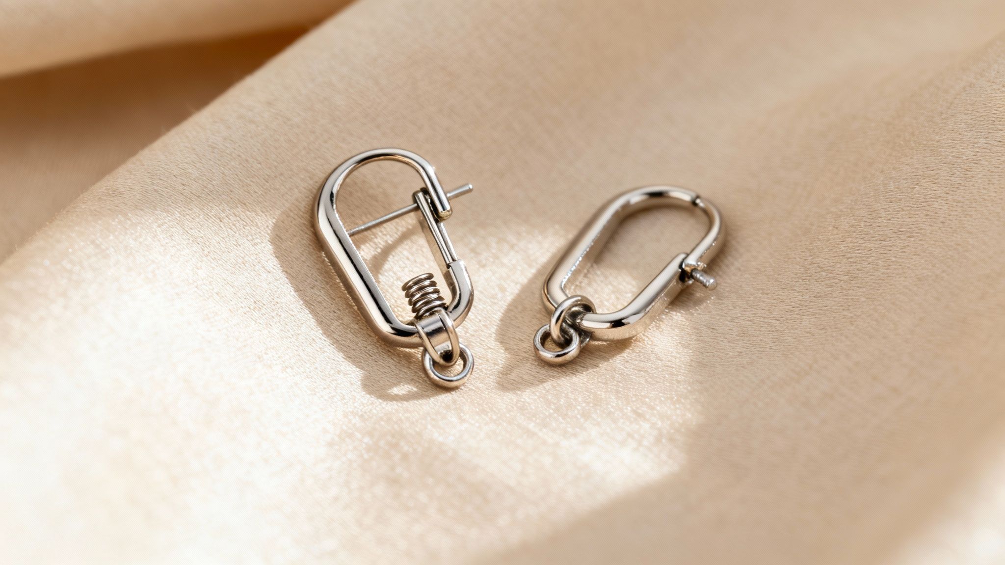 Two silver-colored, oval-shaped earring hooks with spring mechanisms rest on a beige silk fabric.
