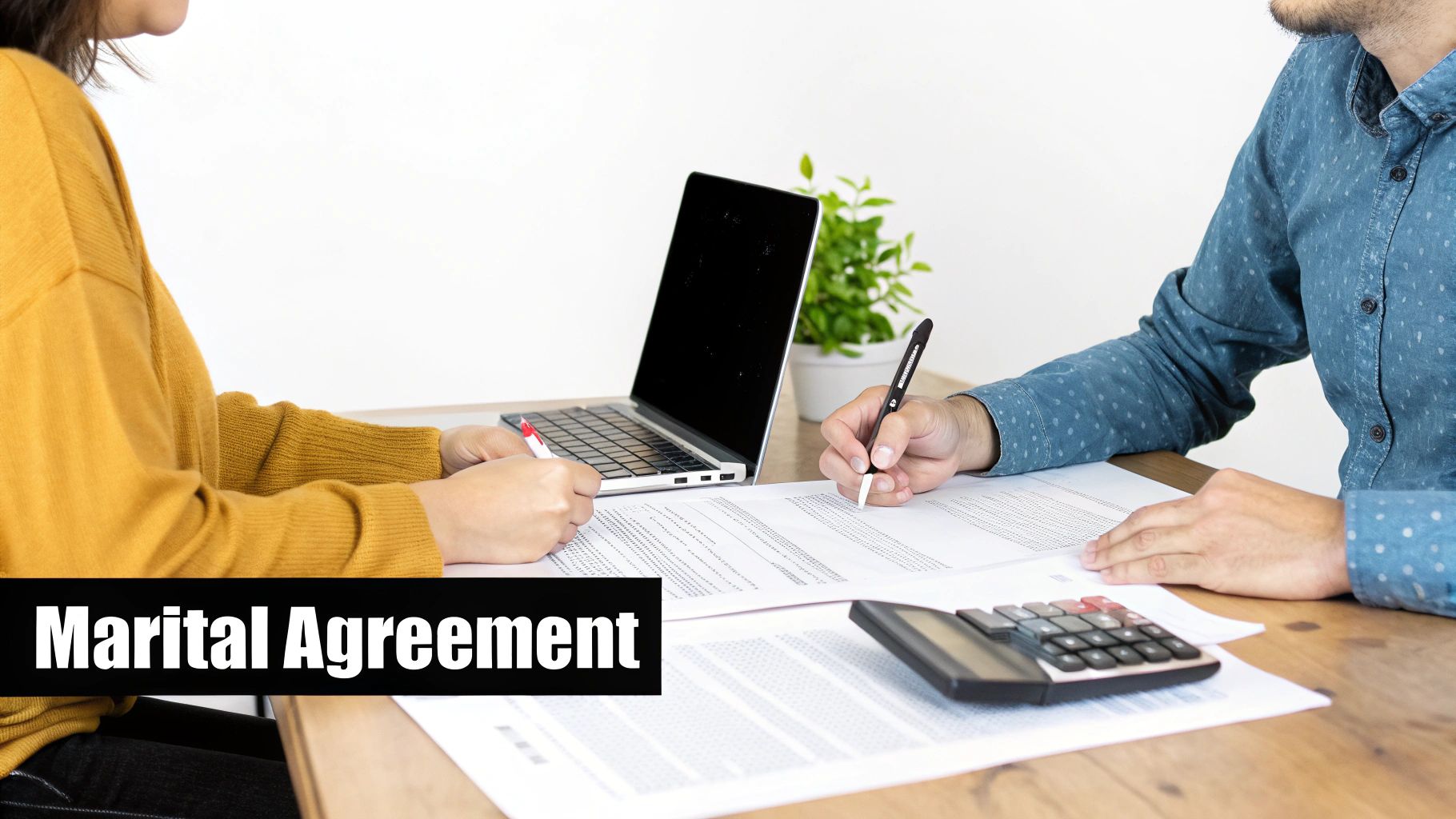 Two people signing a marital agreement document with a laptop and calculator on a wooden table.