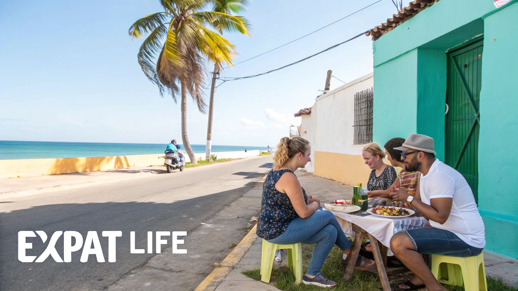 Quatre personnes profitent d'un repas en plein air près d'un bâtiment d'un bleu turquoise éclatant, sur une route côtière ensoleillée.