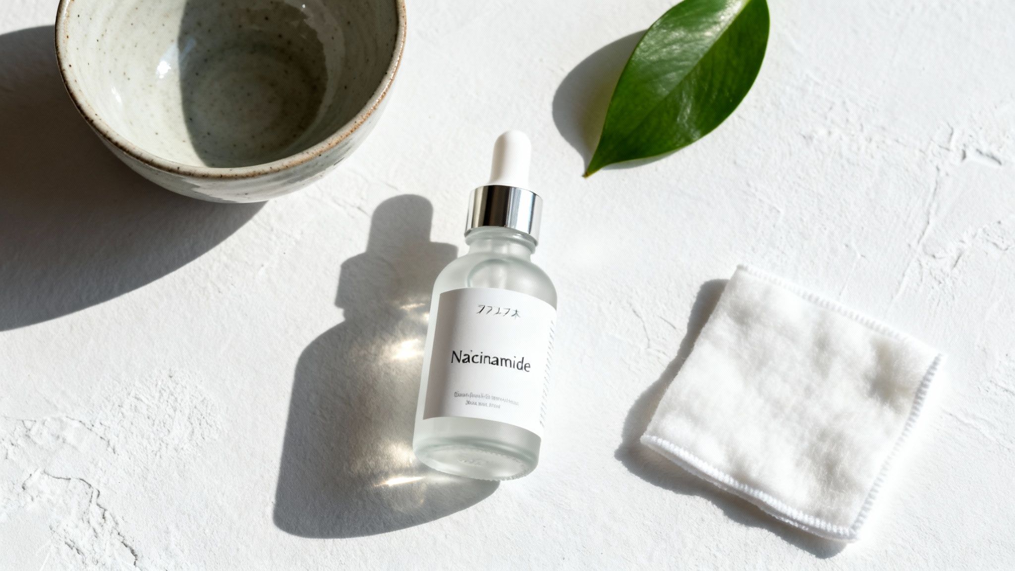 A top-down shot of a niacinamide serum bottle, ceramic bowl, green leaf, and cotton pad on a white surface.