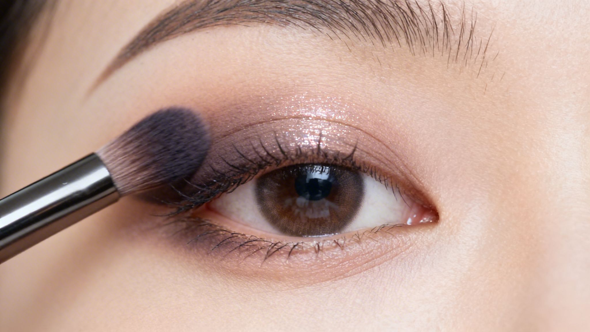 Close-up of an Asian eye receiving smokey, glitter eyeshadow application with a makeup brush.