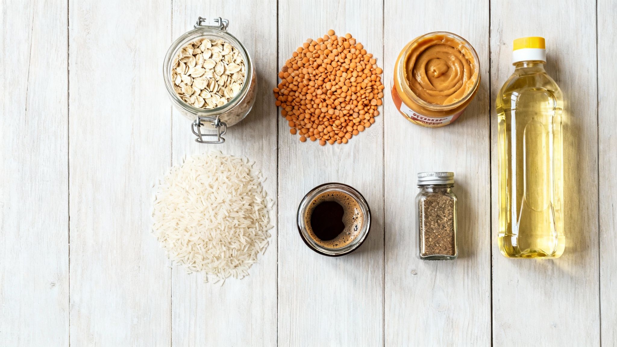Non-perishable food items like oats, lentils, rice, peanut butter, oil, and spices on a white wooden table.