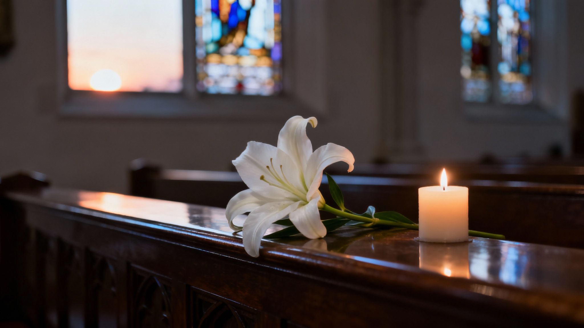 Un lys blanc et une bougie allumée reposent sur un banc d'église en bois, orné de vitraux et d'un coucher de soleil.
