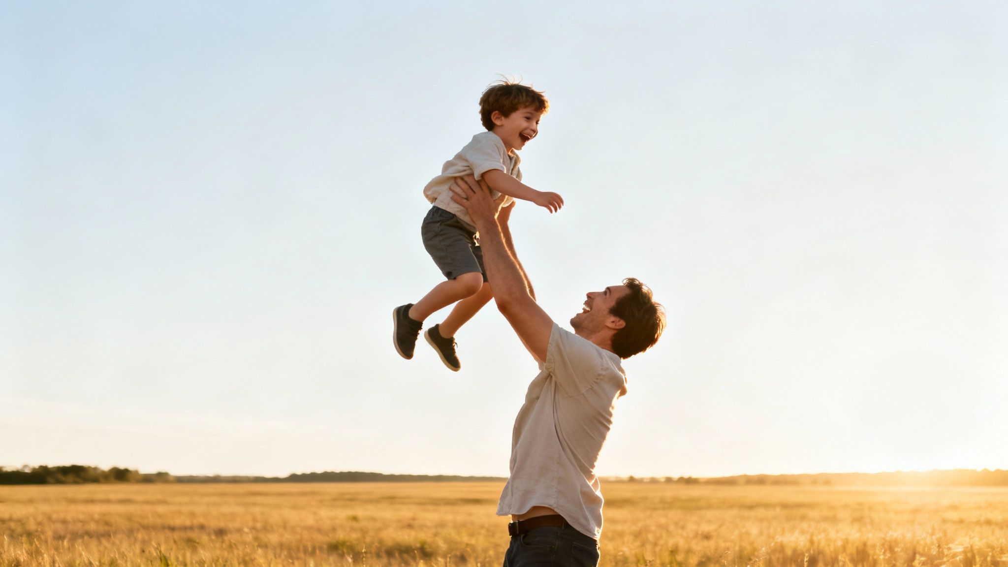 Un père joyeux lance son fils en l'air dans un champ ensoleillé, tous deux riant de bon cœur.