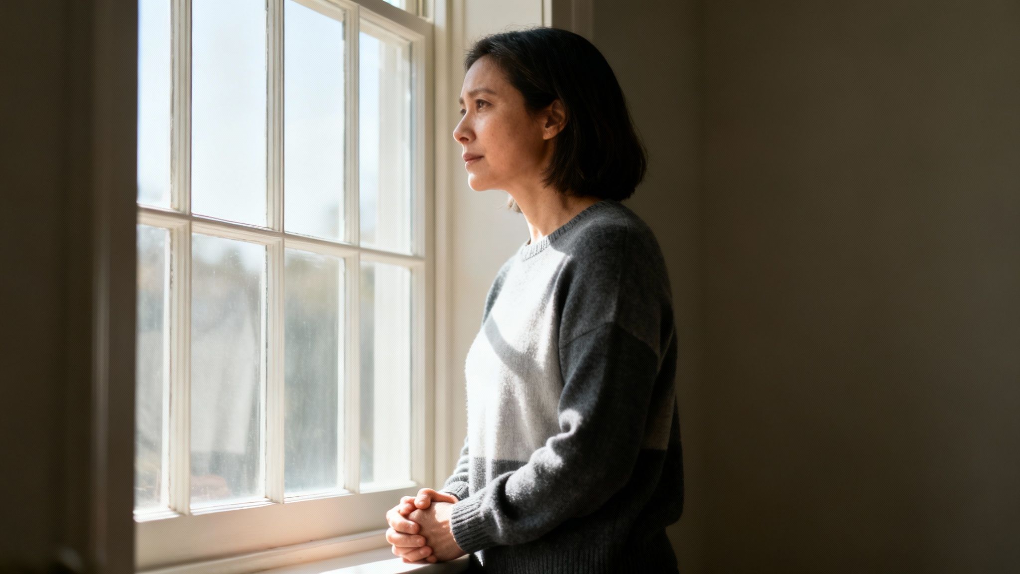 A contemplative woman with dark hair stands by a sunlit window, looking out with a pensive expression.