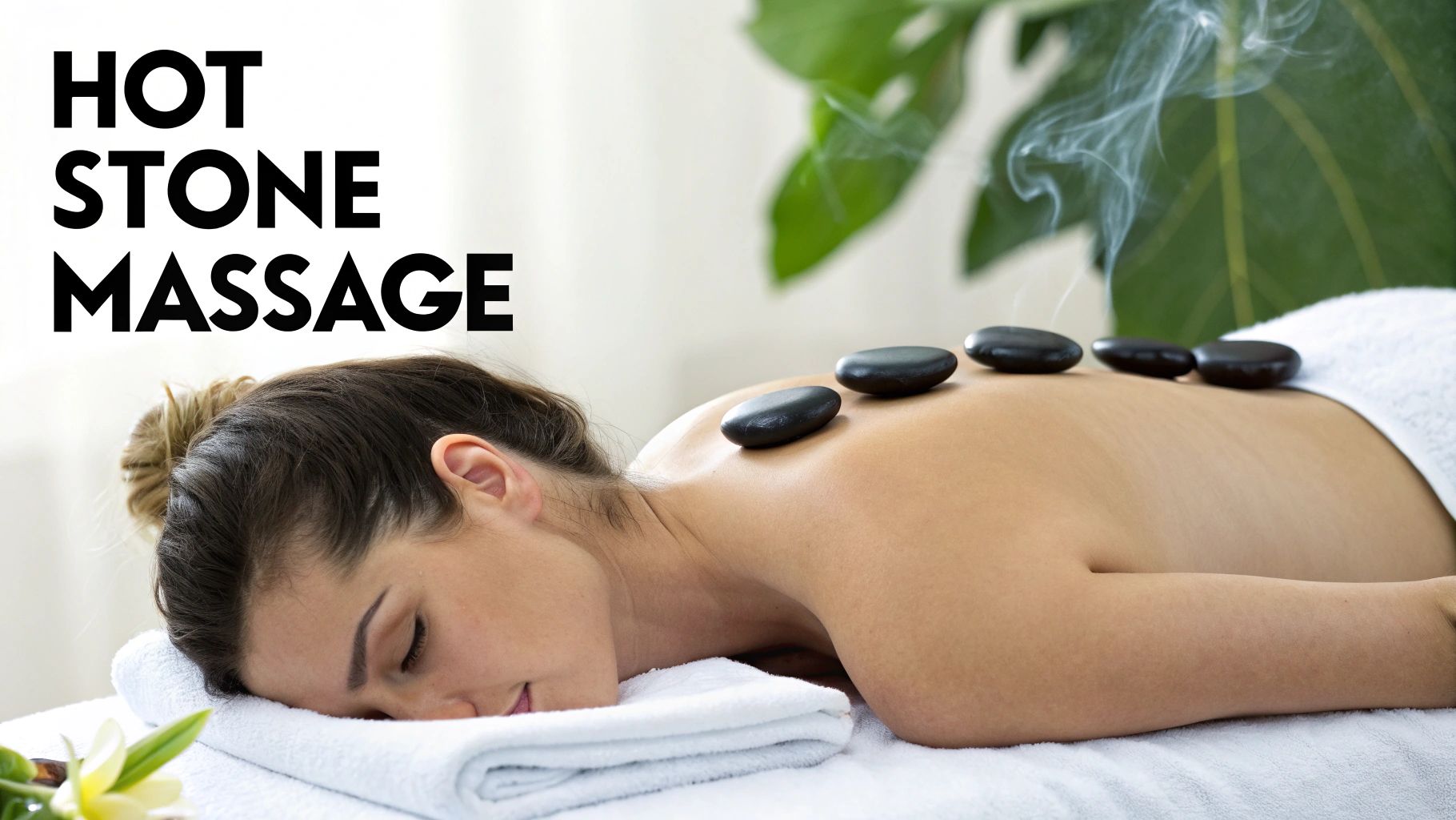 A woman lies relaxed on a white towel, receiving a hot stone massage on her back.