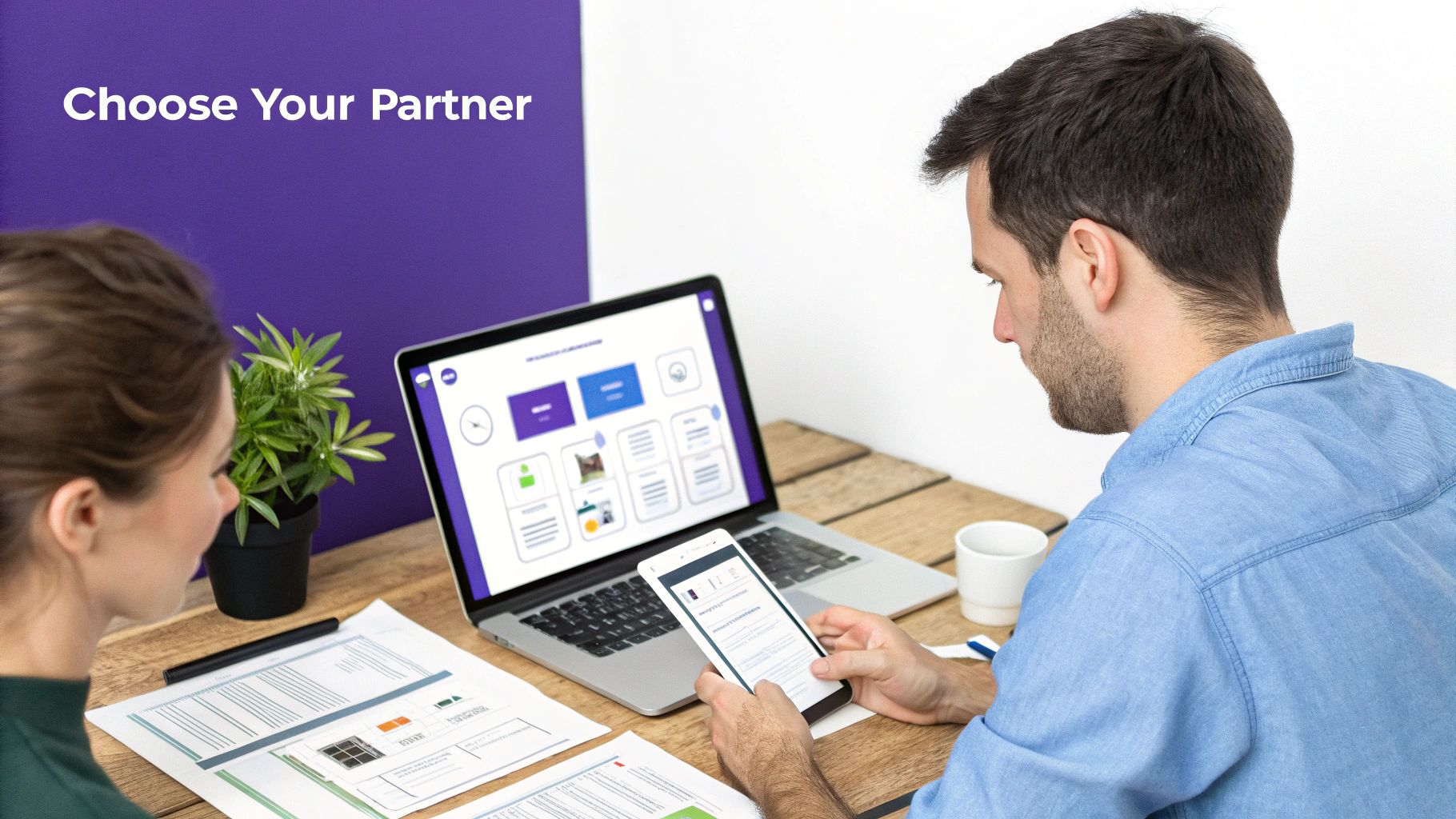 Two people working at a desk with a laptop, smartphone, and documents, likely collaborating on a project.