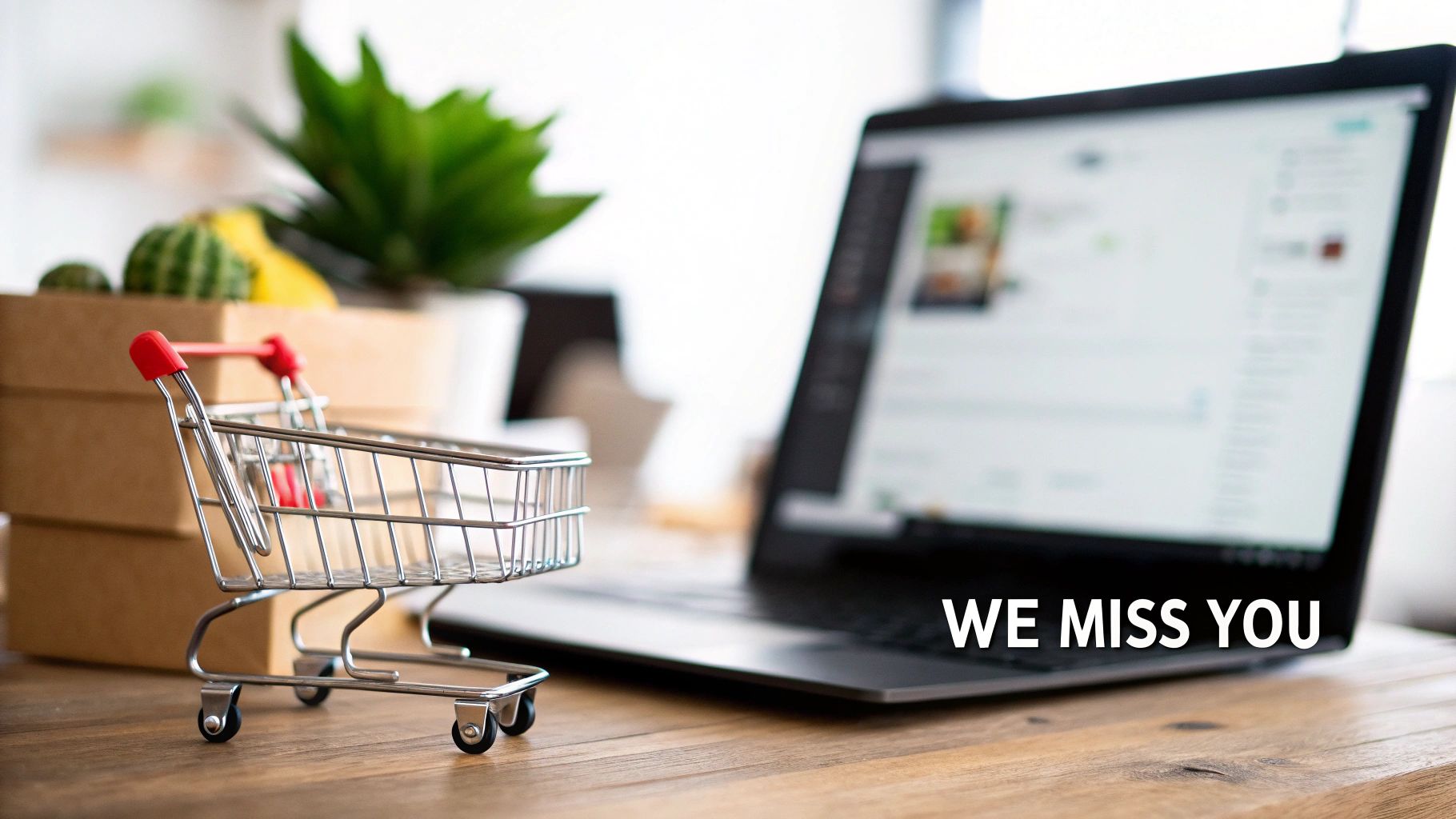 A miniature shopping cart sits on a wooden desk next to a laptop displaying an e-commerce site with 'WE MISS YOU' text.