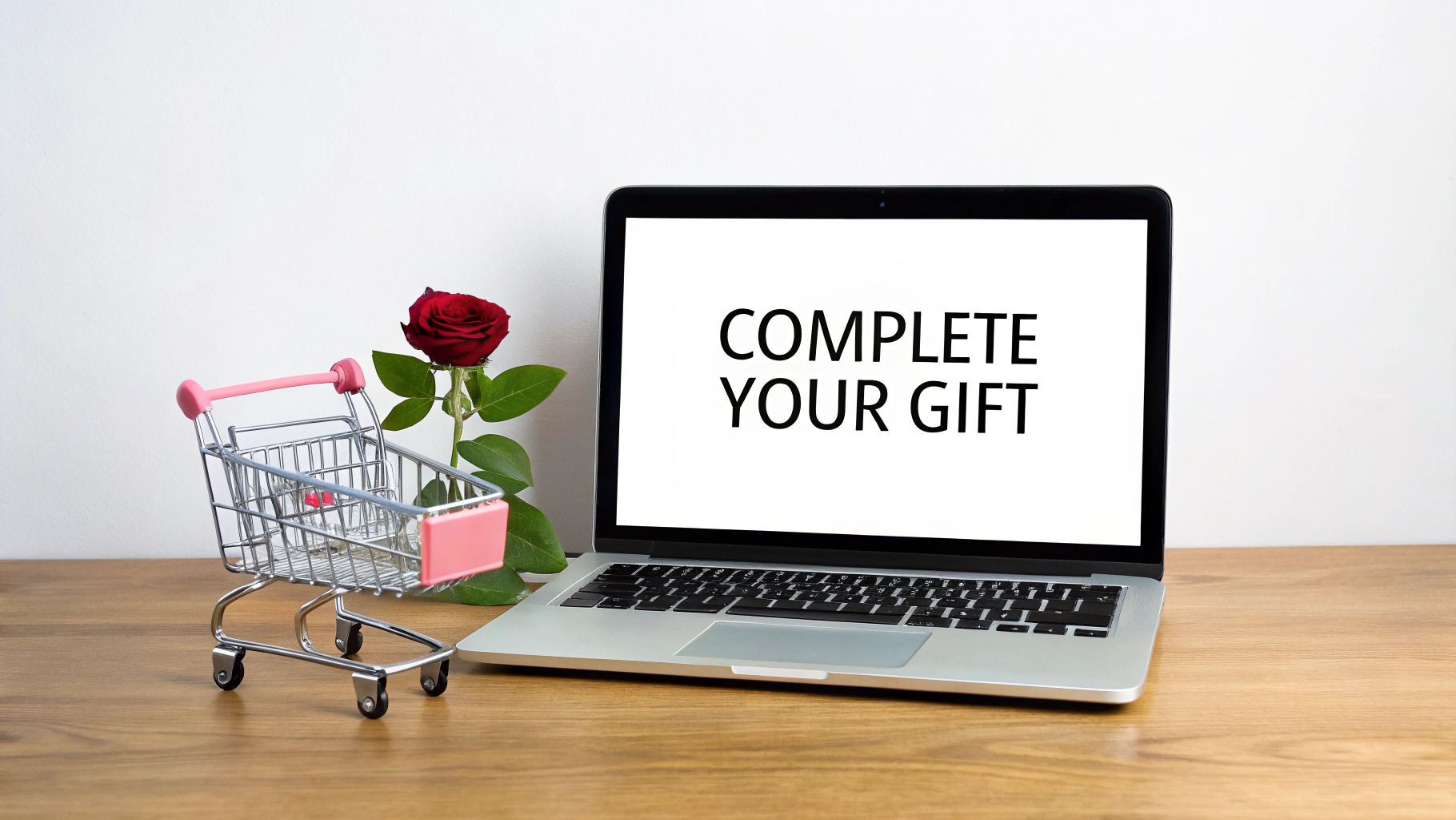 A laptop screen displays 'COMPLETE YOUR GIFT' next to a miniature shopping cart and a red rose on a desk.