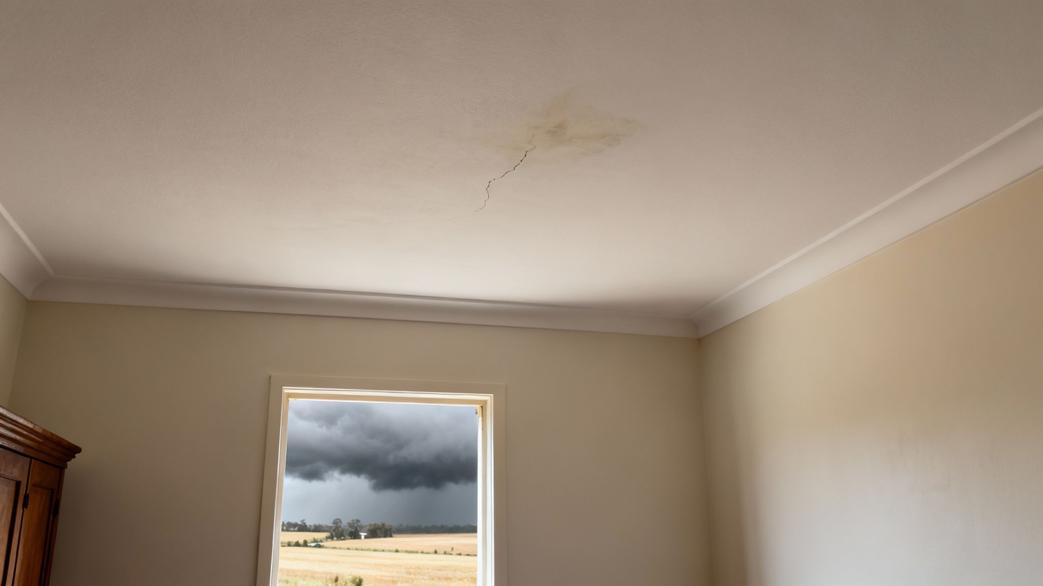 A damaged ceiling with a water stain and crack in a room overlooking a stormy field.