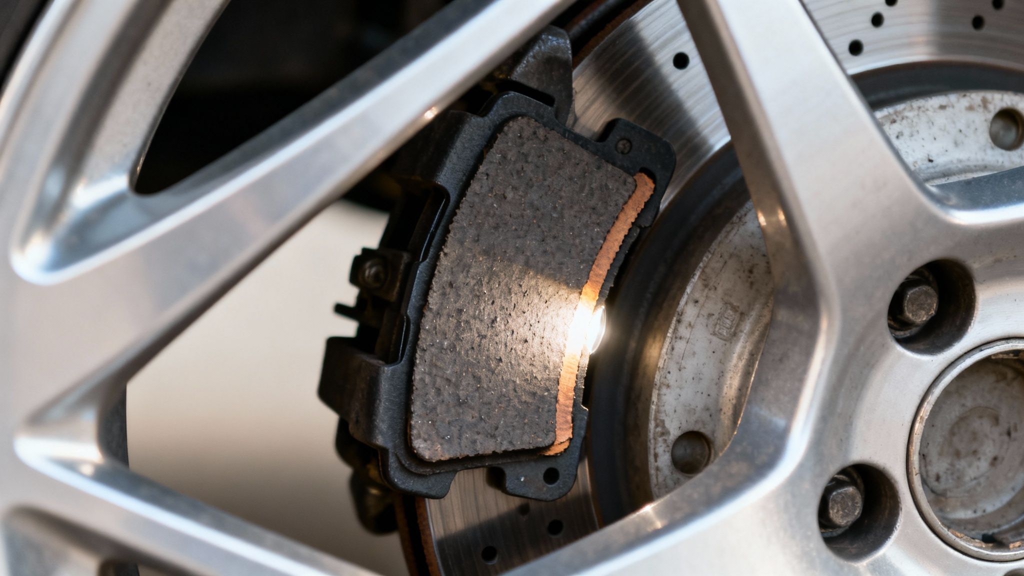 Close-up of a car's worn brake pad and perforated rotor, visible through the silver wheel.