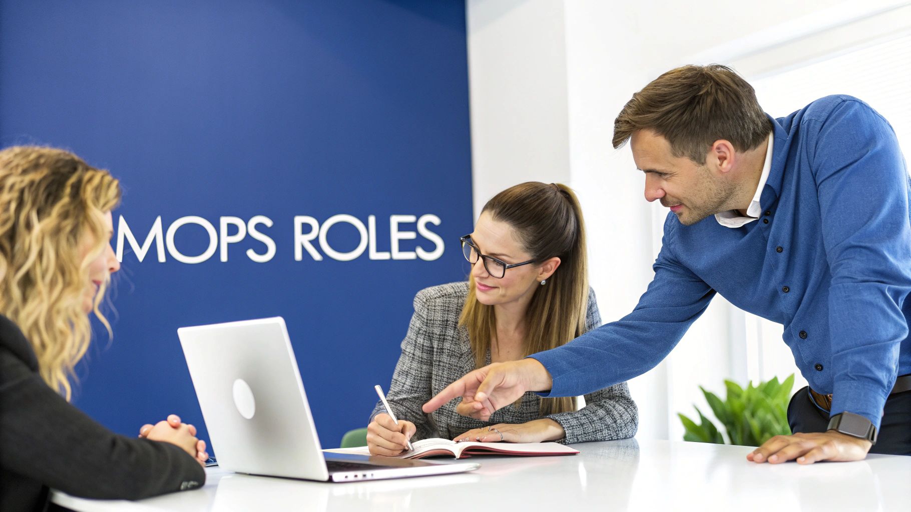 Three diverse professionals collaborating in an office meeting, pointing at a notebook and using a laptop.