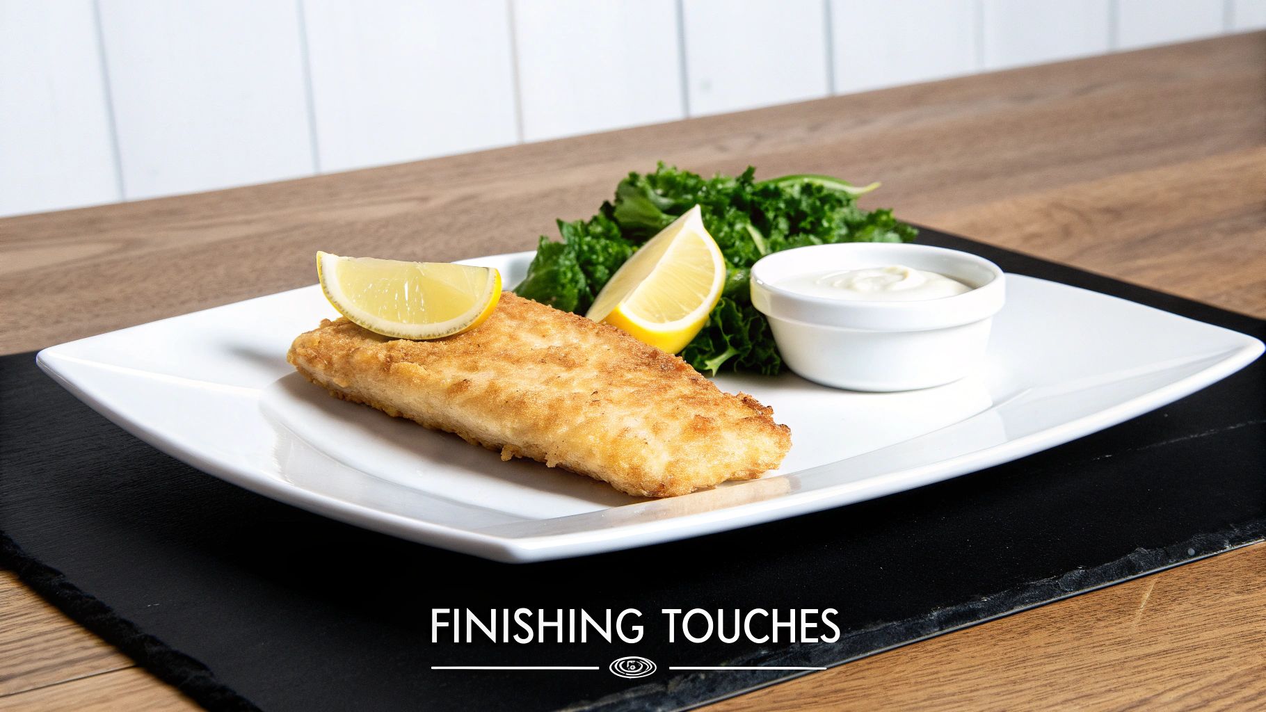 A golden-brown fried fish fillet served with fresh lemon wedges, green kale, and a side of white sauce on a plate.
