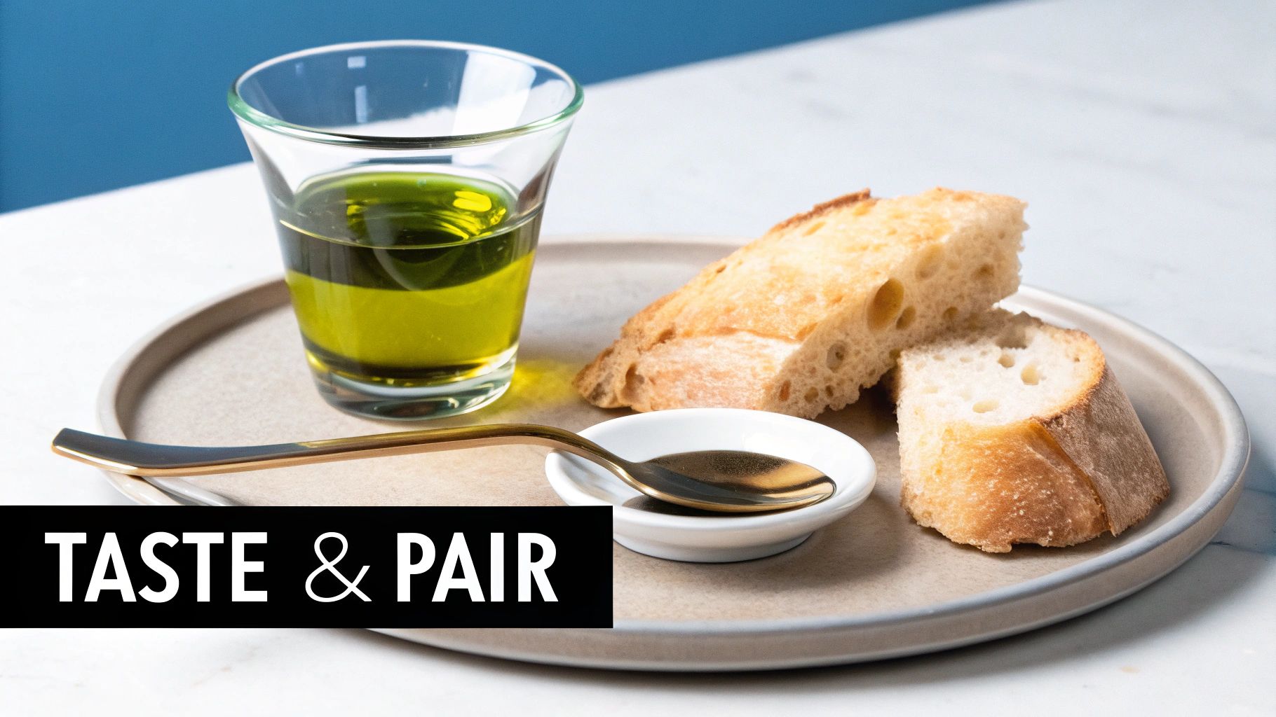A glass of golden olive oil, fresh bread slices, a spoon, and a small dish on a beige plate.