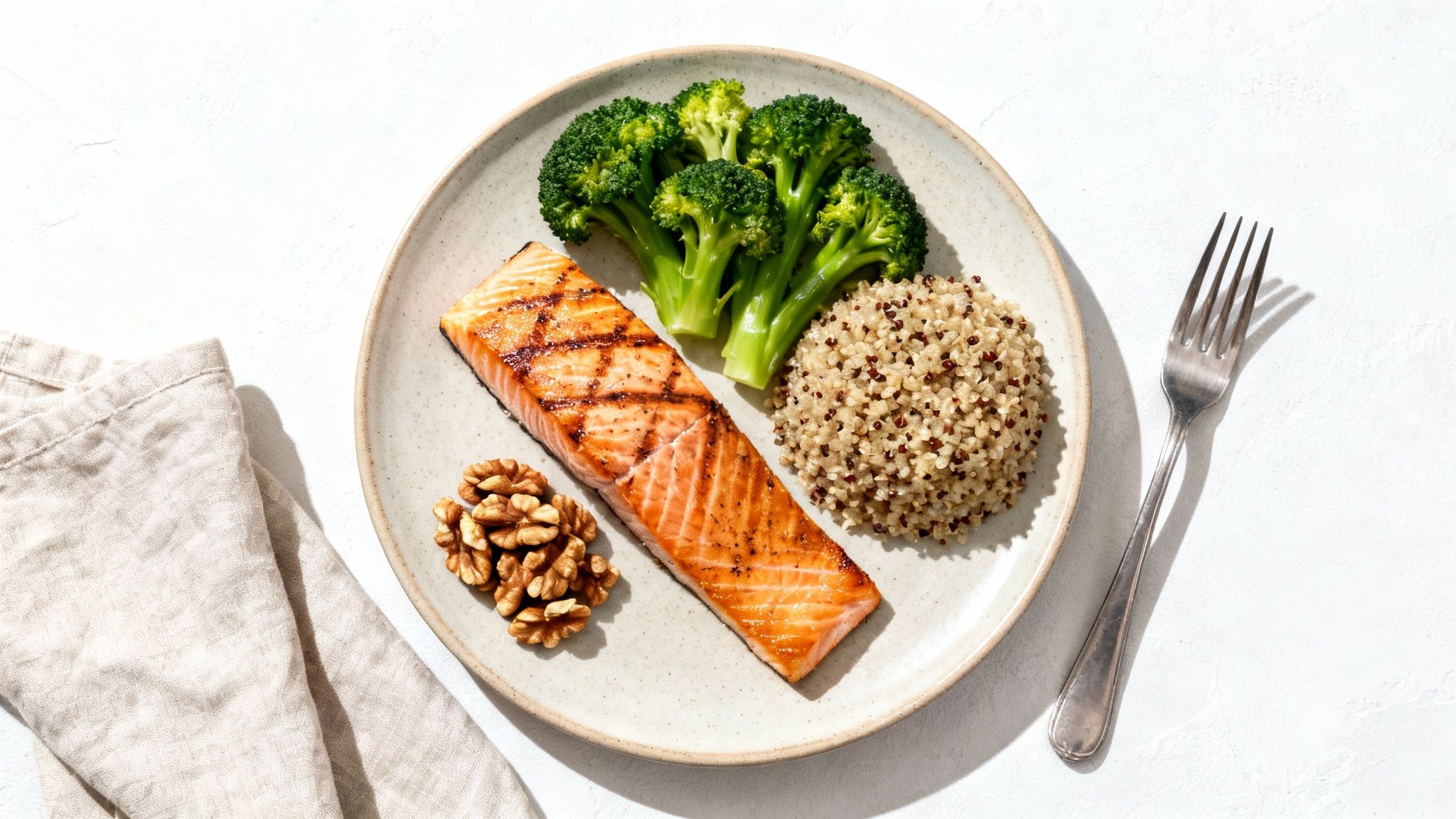 A healthy meal with grilled salmon, steamed broccoli, quinoa, and walnuts on a plate, with a fork and napkin.