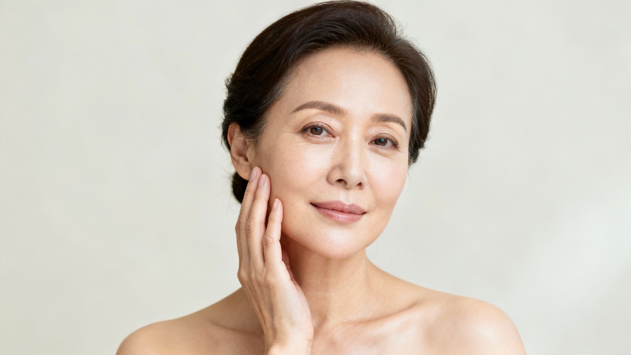 An elegant senior Asian woman with clear, radiant skin smiles, gently touching her cheek.