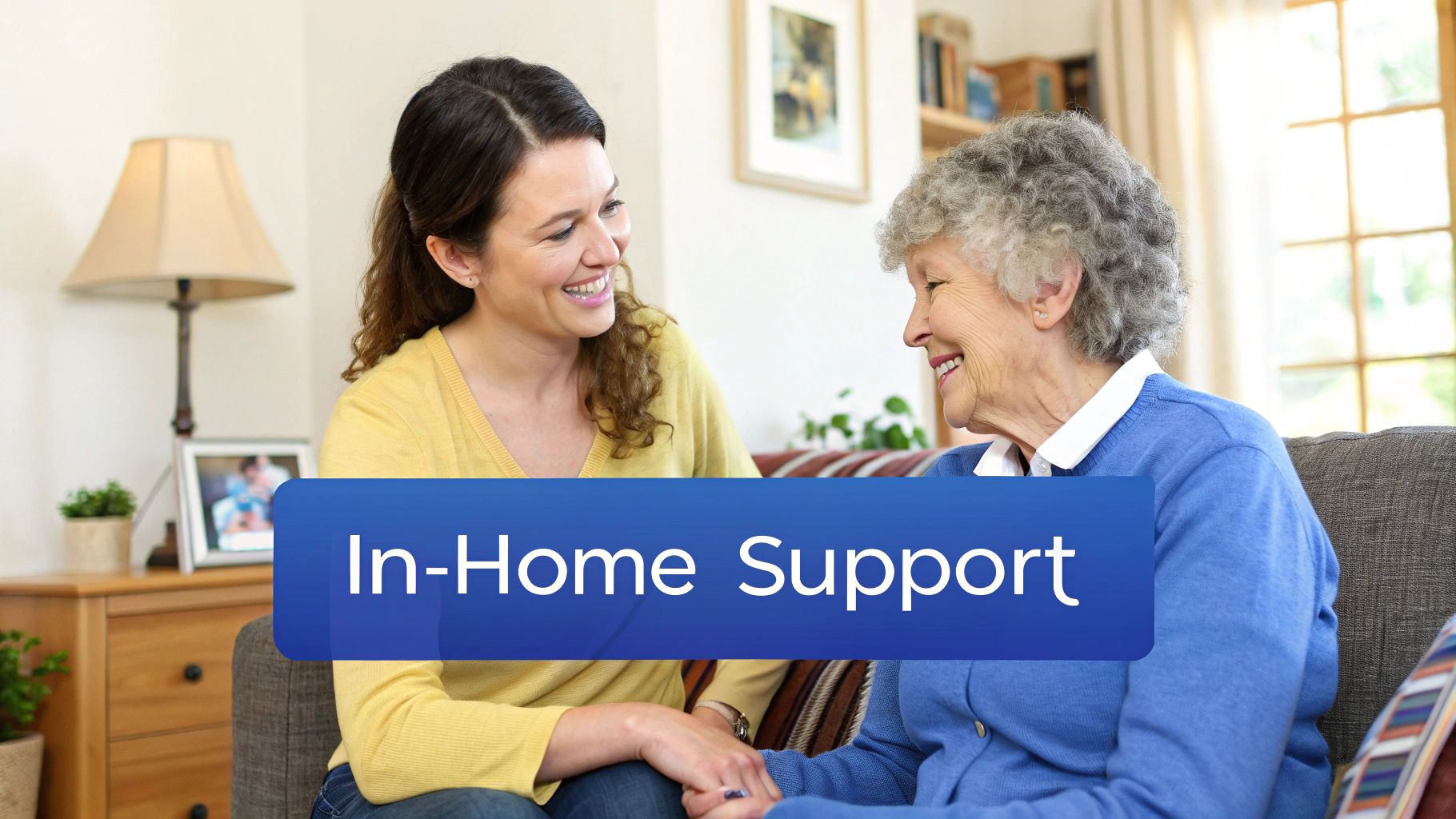 A smiling young woman holding the hand of a happy older woman on a couch, representing in-home support.