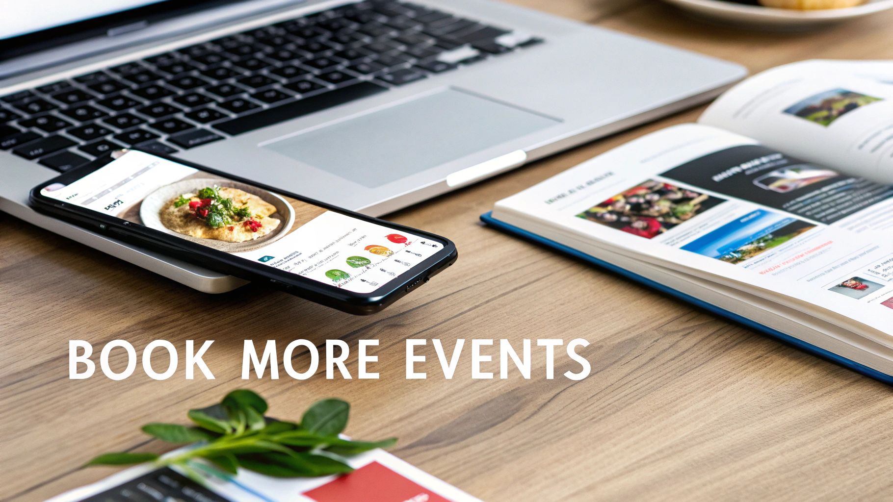 A laptop, smartphone displaying a food image, and an open book on a wooden desk.