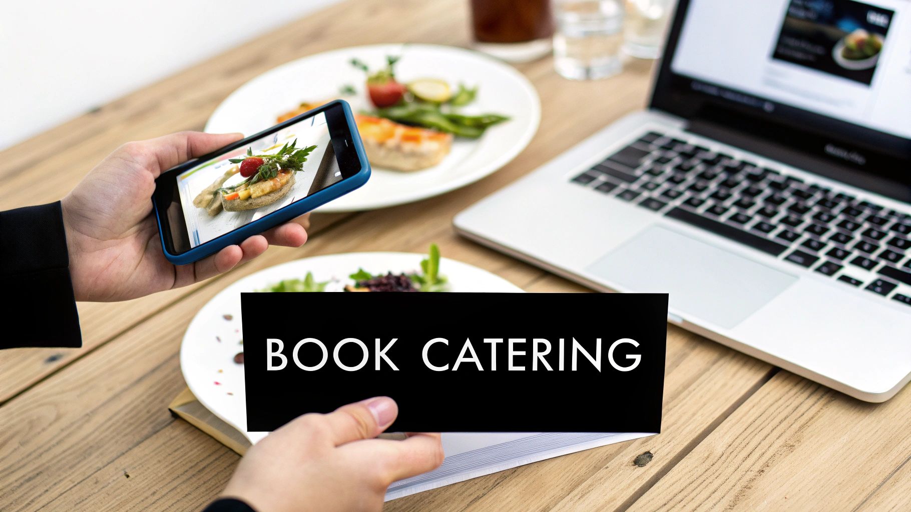 Person holds smartphone taking photo of gourmet food, with a laptop and 'BOOK CATERING' sign on a wooden table.