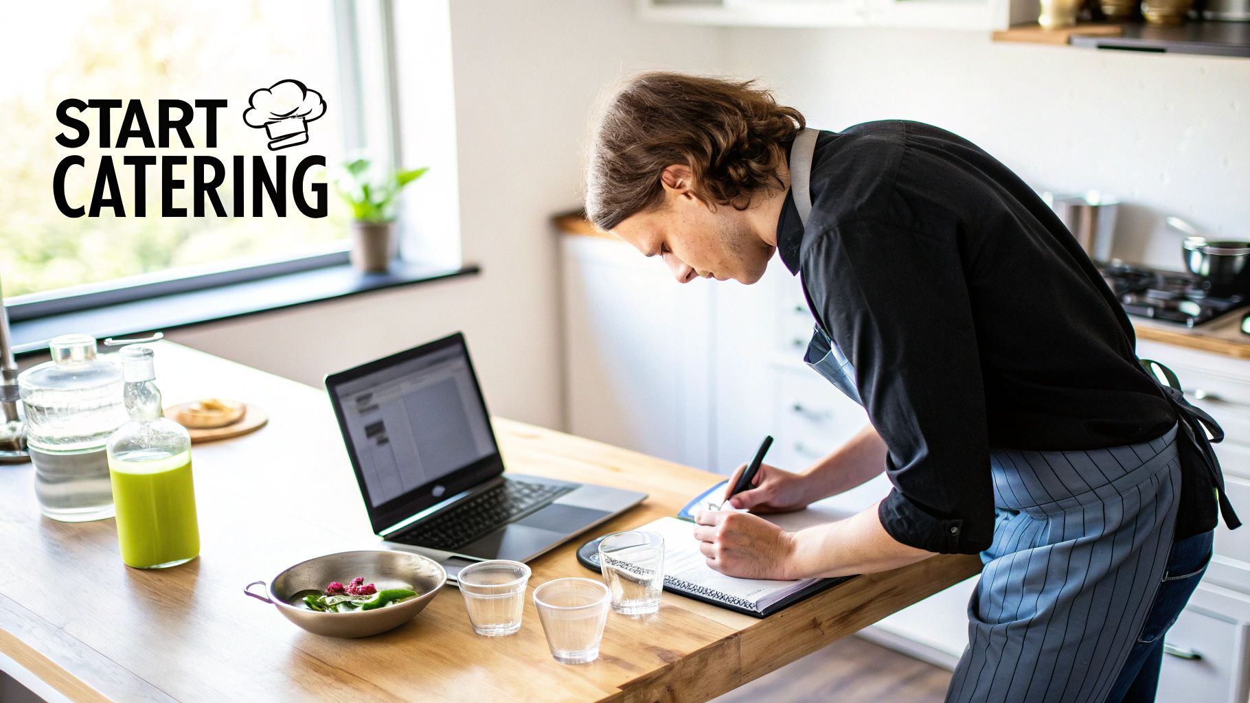 A man in an apron writing at a kitchen counter with a laptop, juice, and food, planning a catering business.