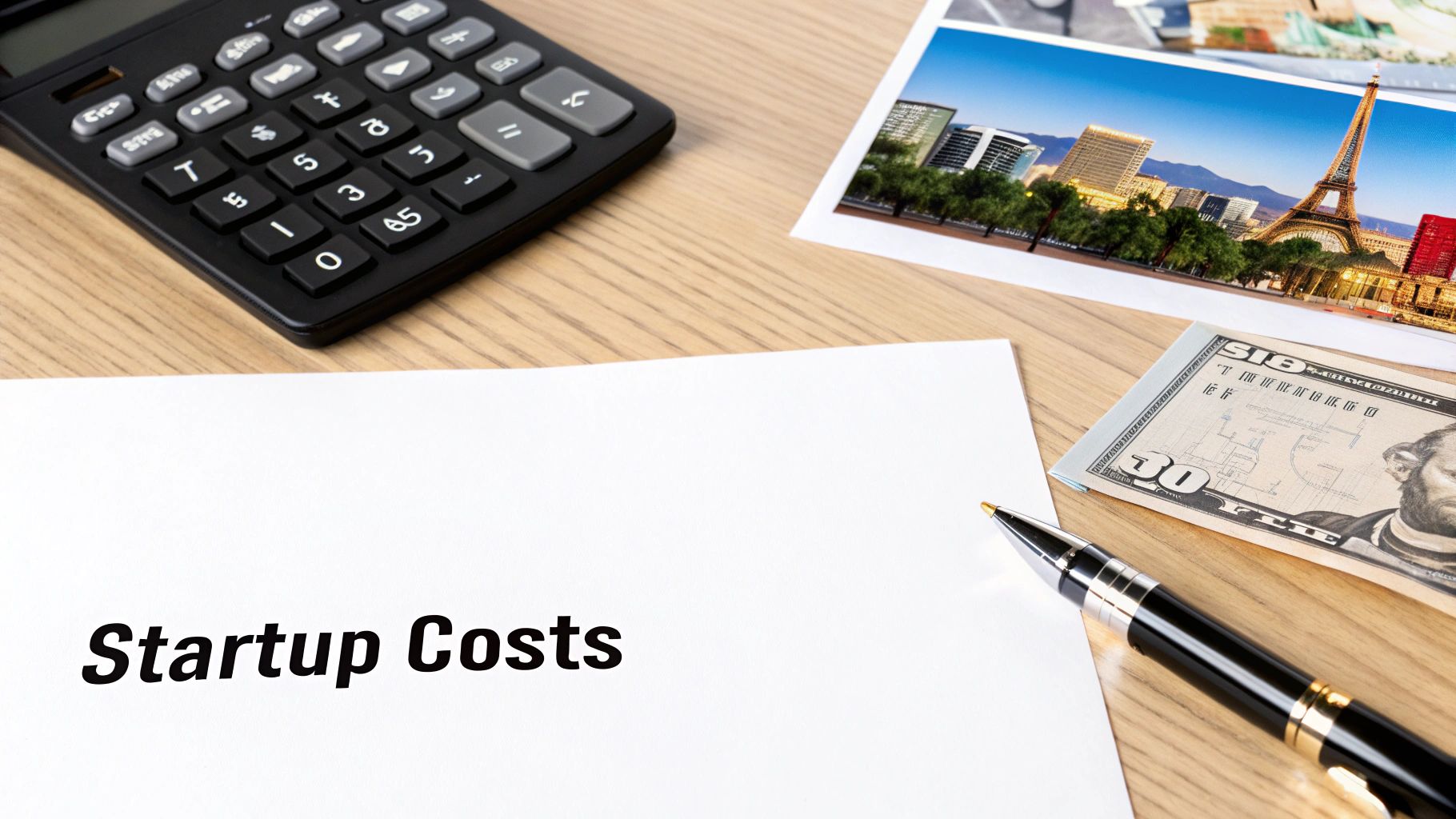 A desk setup with a calculator, paper marked 'Startup Costs', a pen, money, and a city photo.
