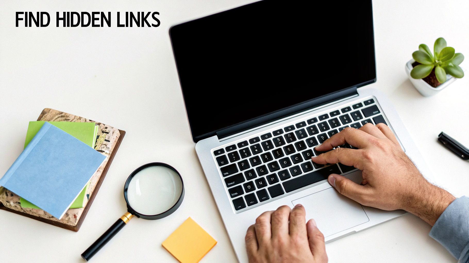 Hands typing on a silver laptop next to a magnifying glass, notebooks, and a plant.