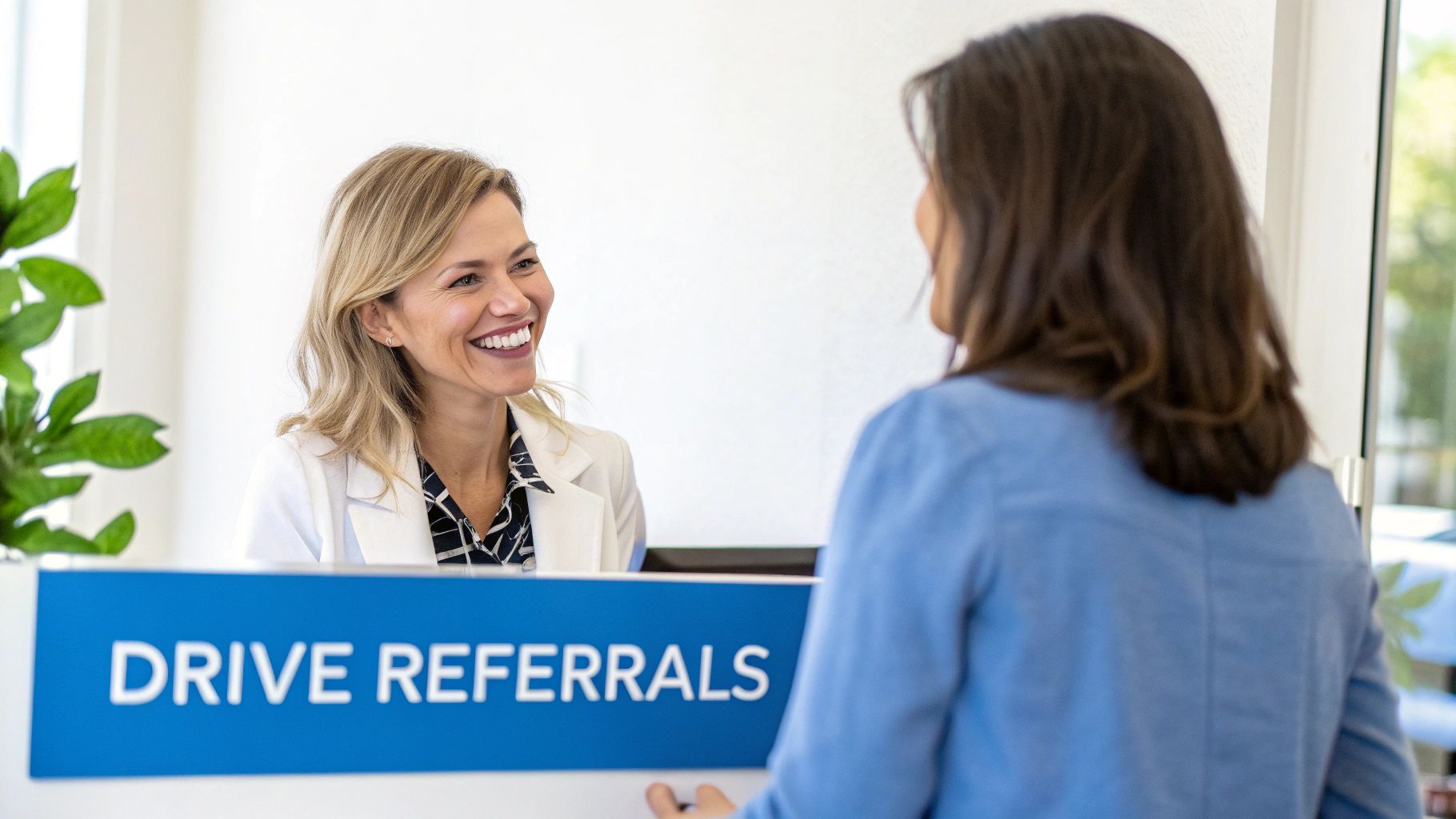 A smiling woman at a clinic front desk talks to a client under a "DRIVE REFERRALS" sign.