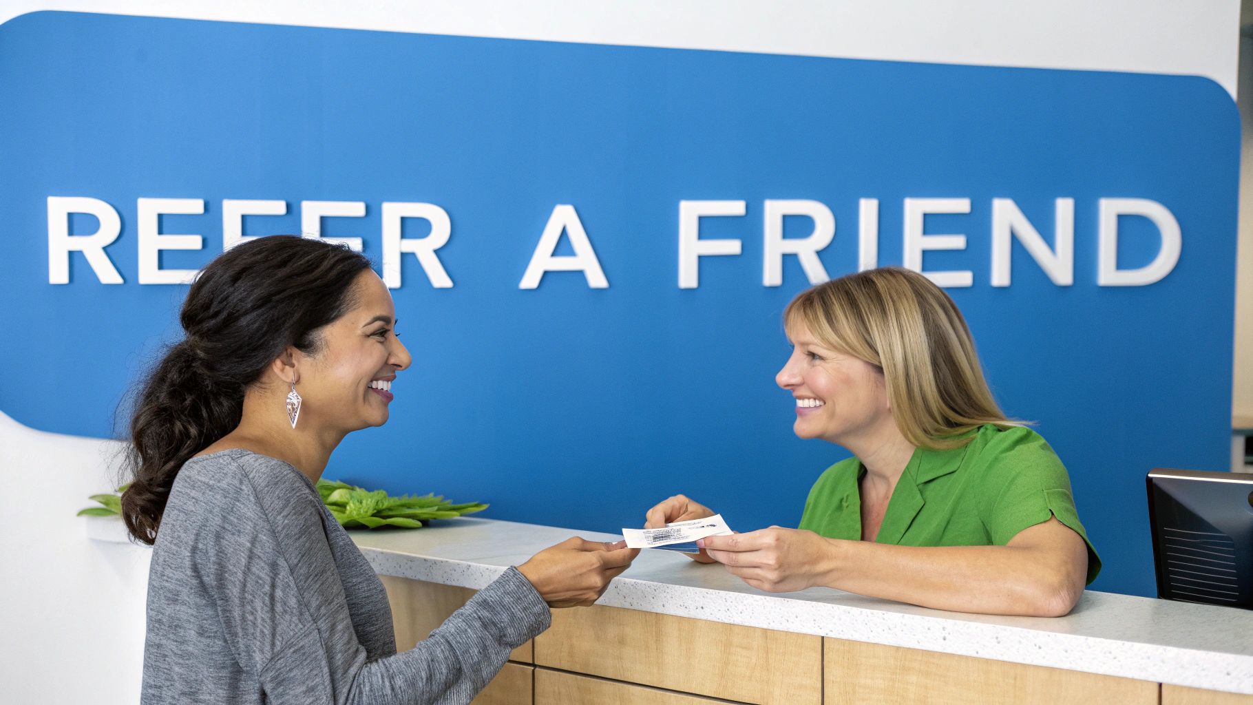 Two smiling women exchange a refer-a-friend card at a reception desk with a blue sign.