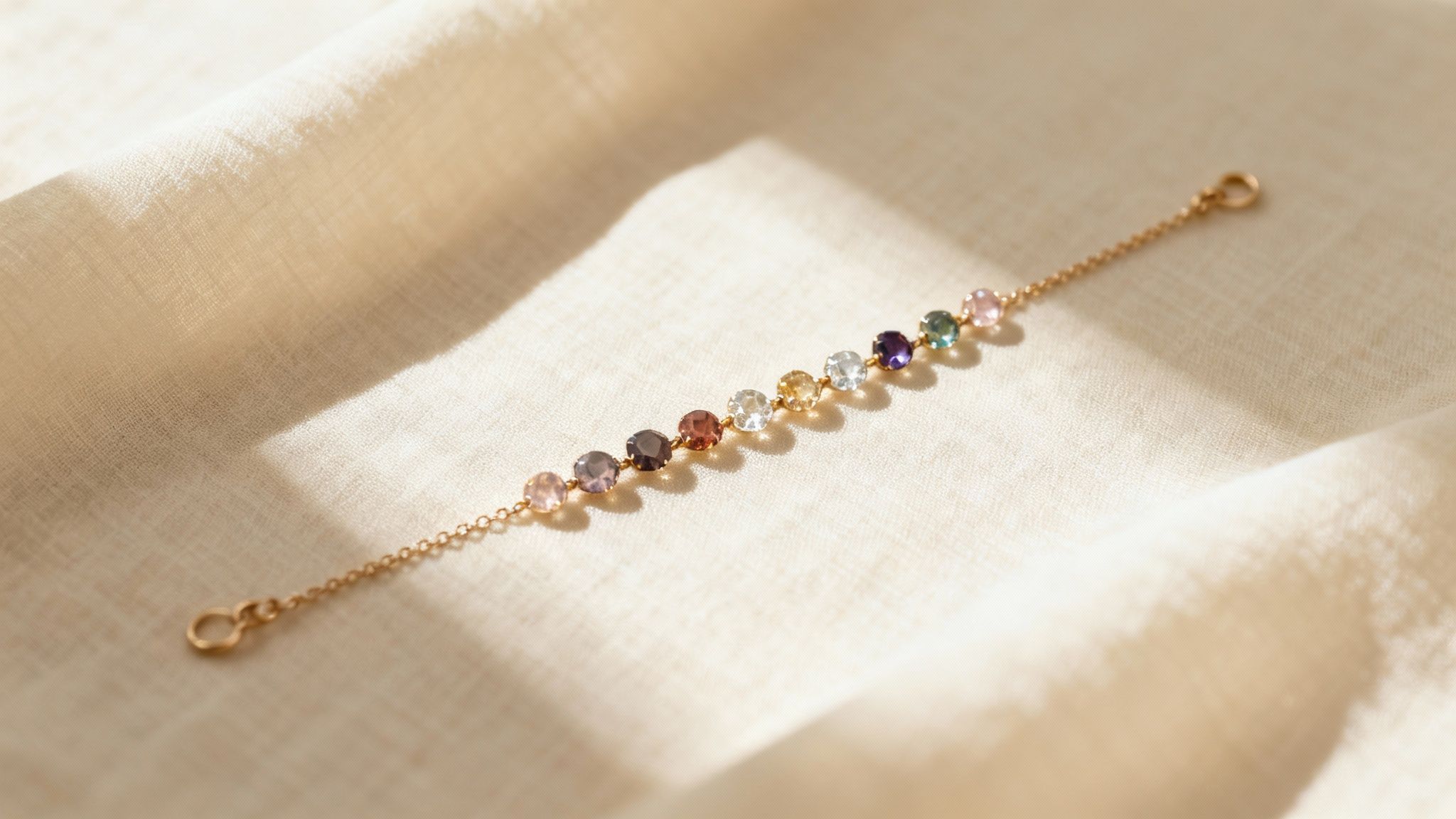 A delicate gold chain bracelet featuring a row of colorful round gemstones rests on a light fabric.