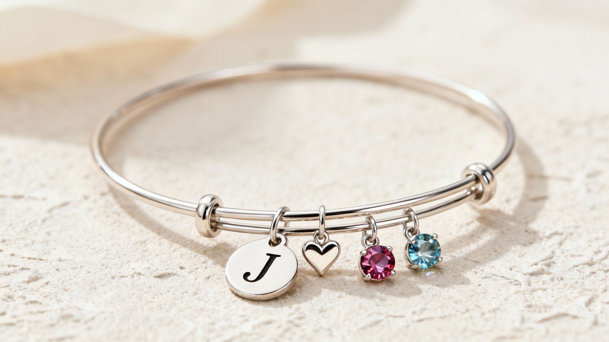 A silver adjustable bangle bracelet featuring a 'J' initial, a heart, and two colorful birthstone charms.
