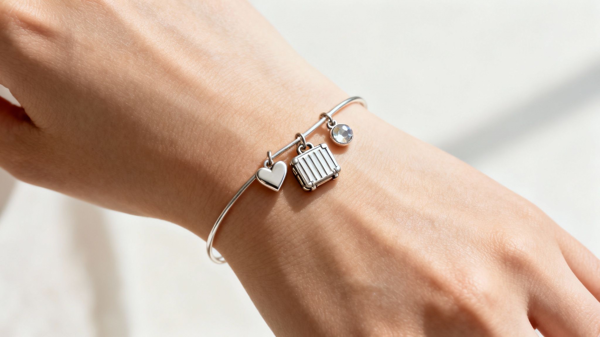 Close-up of a hand wearing a silver charm bracelet with heart, suitcase, and gemstone.