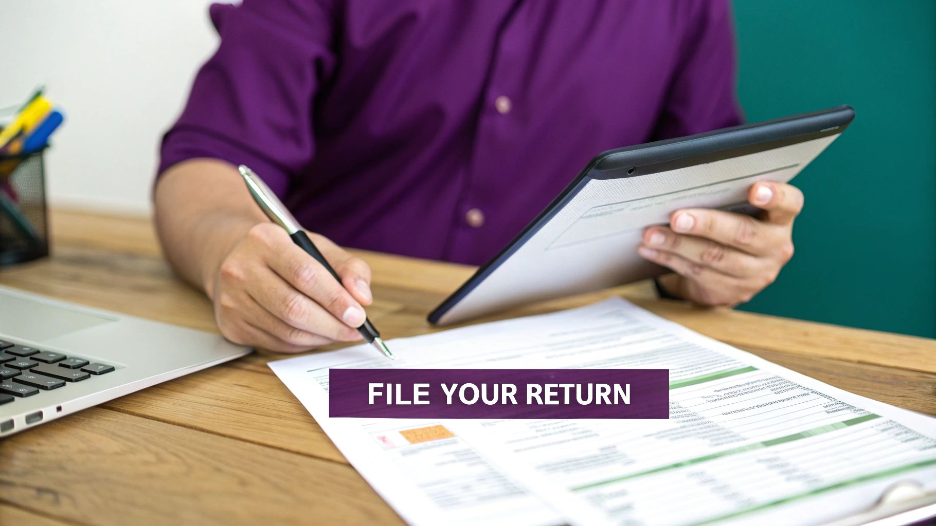 Person preparing to file tax returns, writing on documents with a pen, laptop and tablet on desk.